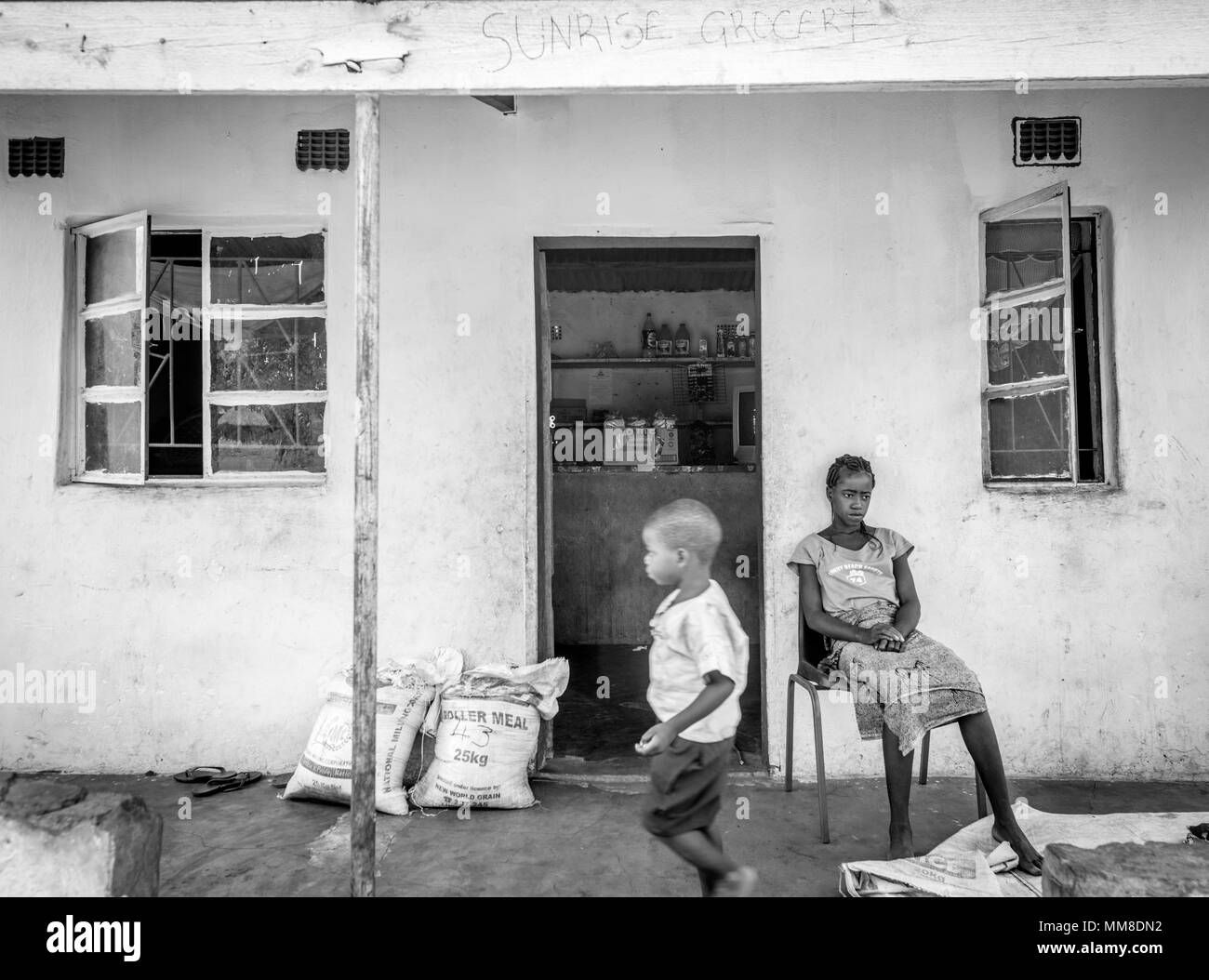 Young Zambian woman sits in chair placed out in front of village's local grocery store as a young boy runs past her, Mukuni Village, Zambia Stock Photo