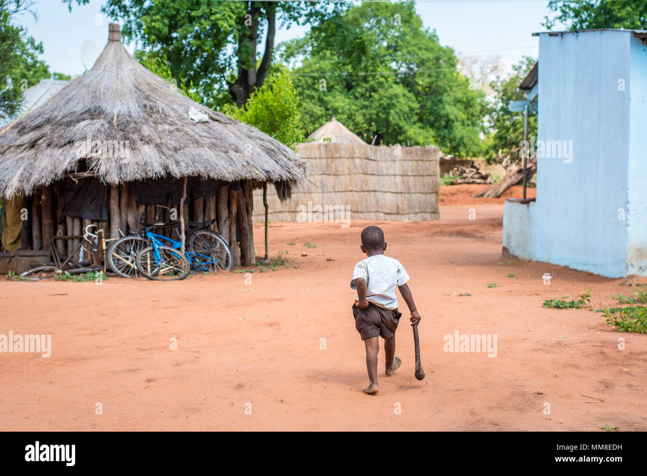 Young Zambian boy walks away through village while holding onto hatchet and his pants, Mukuni Village, Zambia Stock Photo