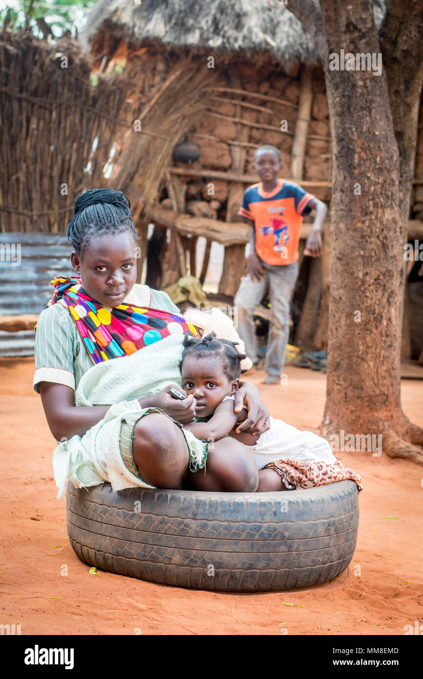 Young Zambian mother sits in tire with her toddler by her side and baby in sling, Mukuni Village, Zambia Stock Photo