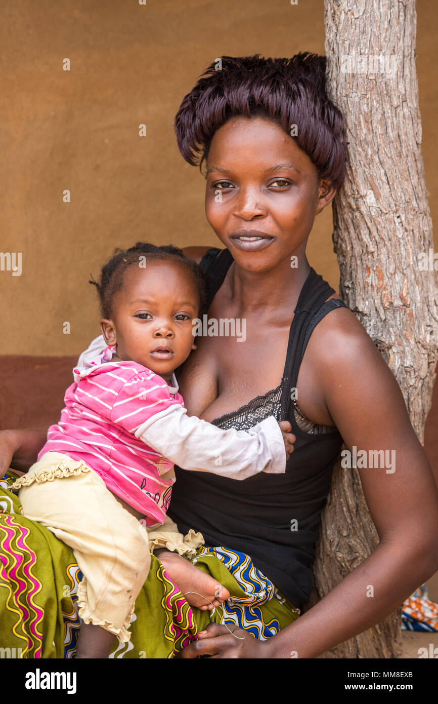 Young Zambian mother leans against tree post with her baby sitting in her lap, Mukuni Village, Zambia Stock Photo