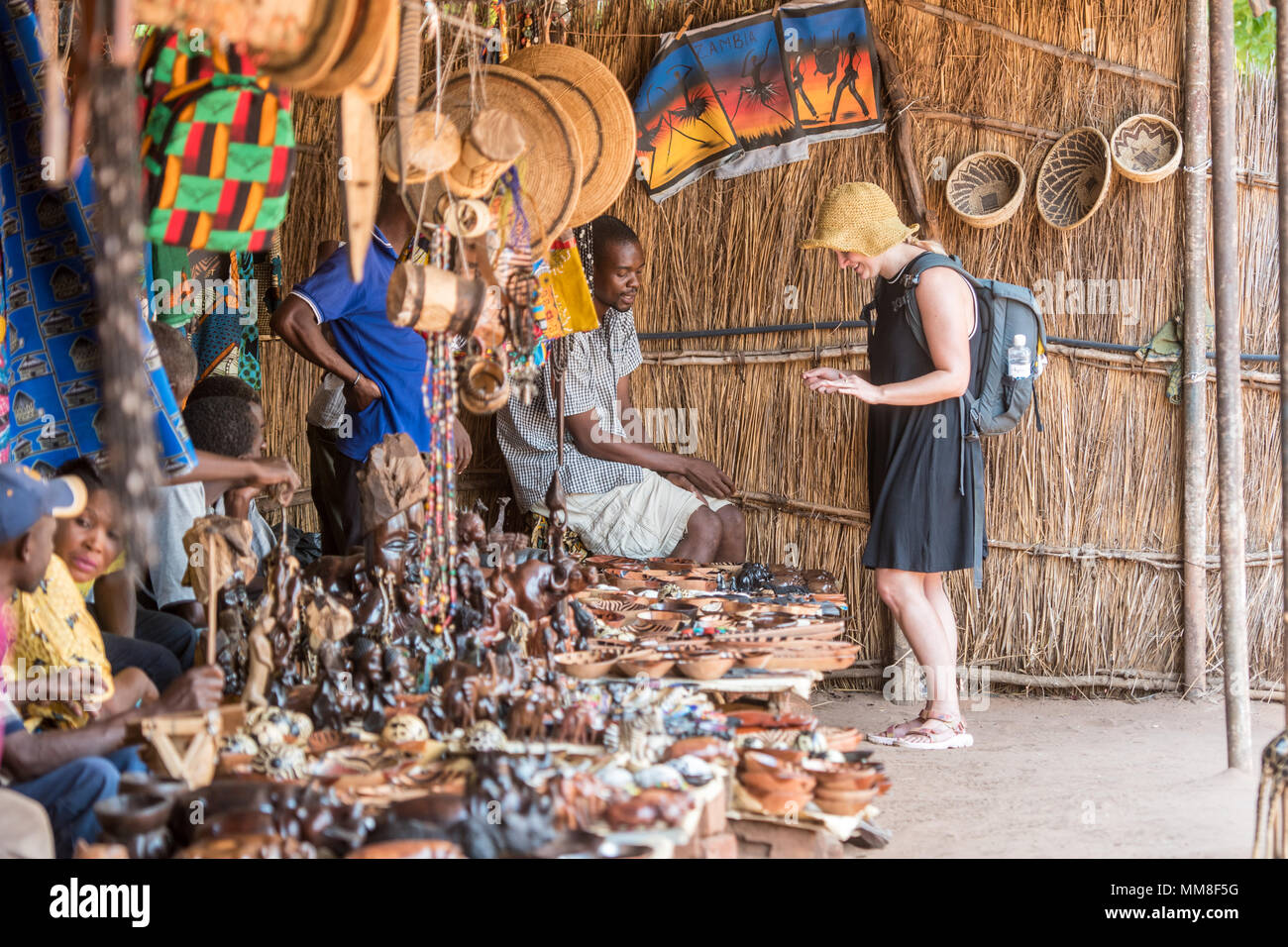 Young woman tourist browses over table full of handcrafted souvenirs while vendor assists her with purchase, Mukuni Village, Zambia Stock Photo