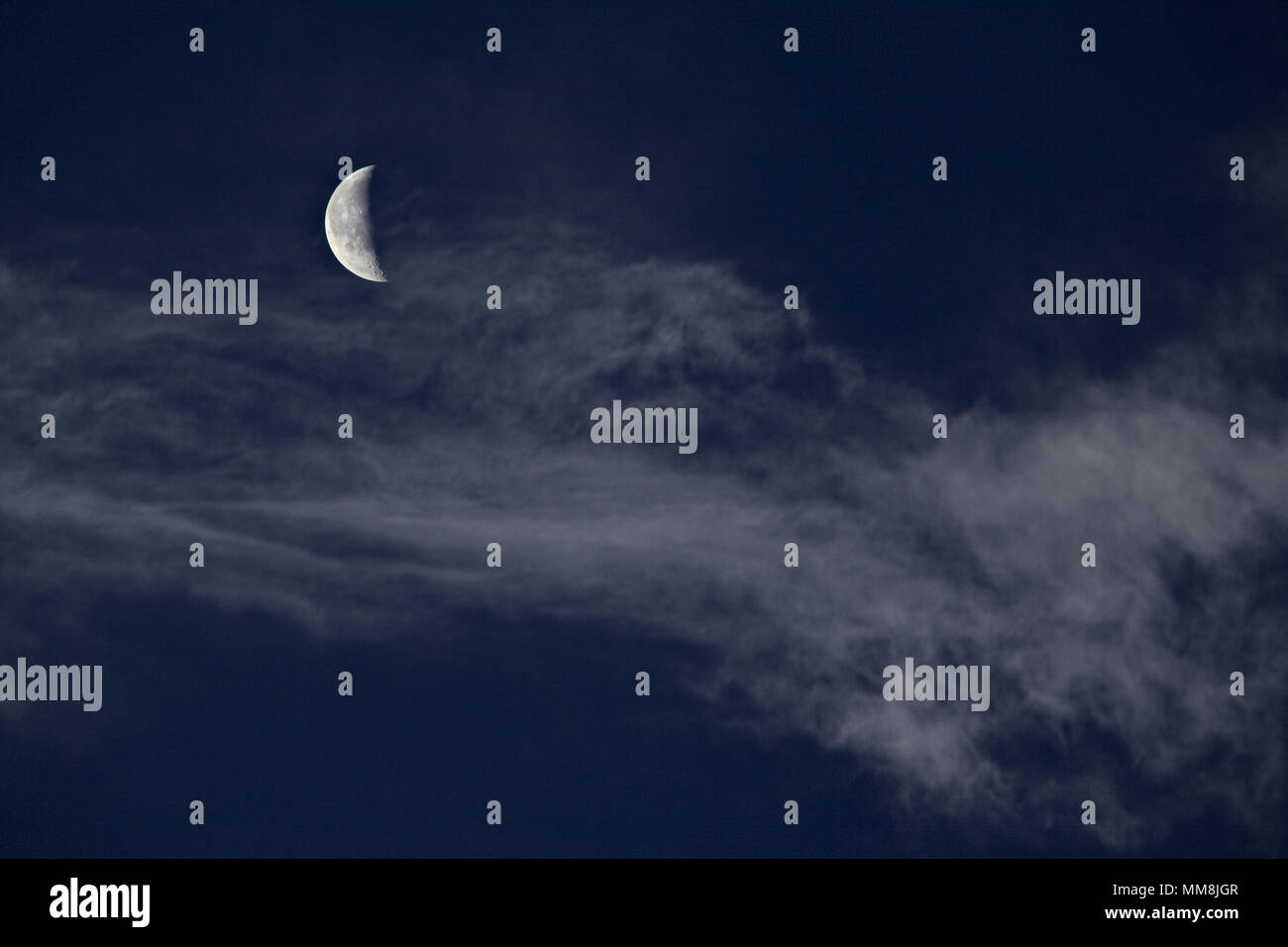 Crescent moon behind high altitude cirrus clouds against a blue sky Stock Photo