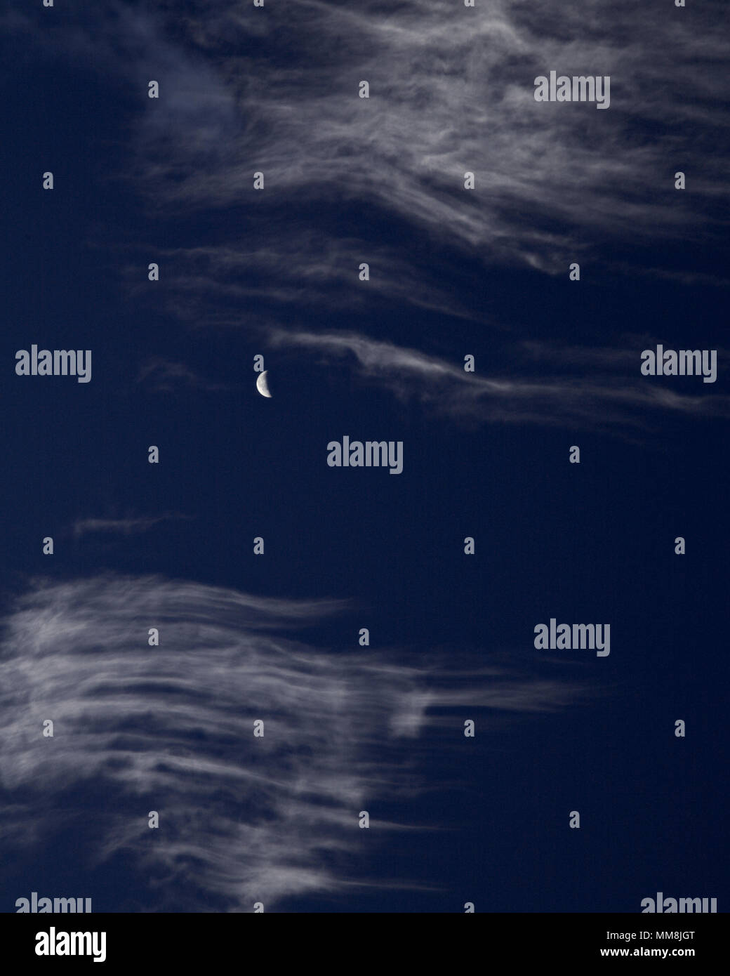Crescent moon behind high altitude cirrus clouds against a blue sky Stock Photo