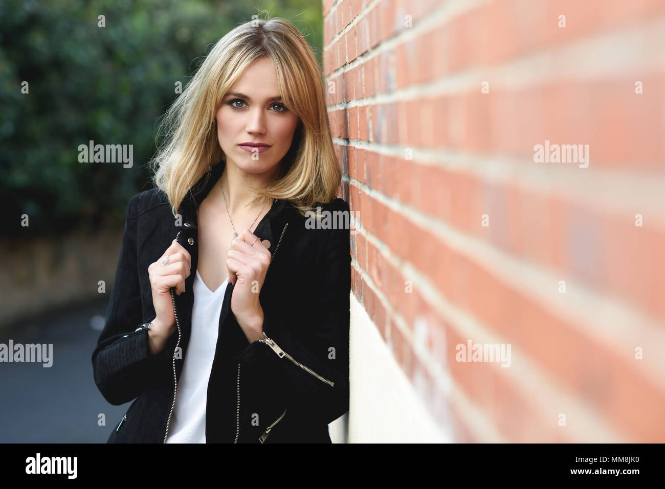Attractive blonde woman in urban background. Young girl wearing black zipper jacket standing in the street. Pretty female with straight hair hairstyle Stock Photo
