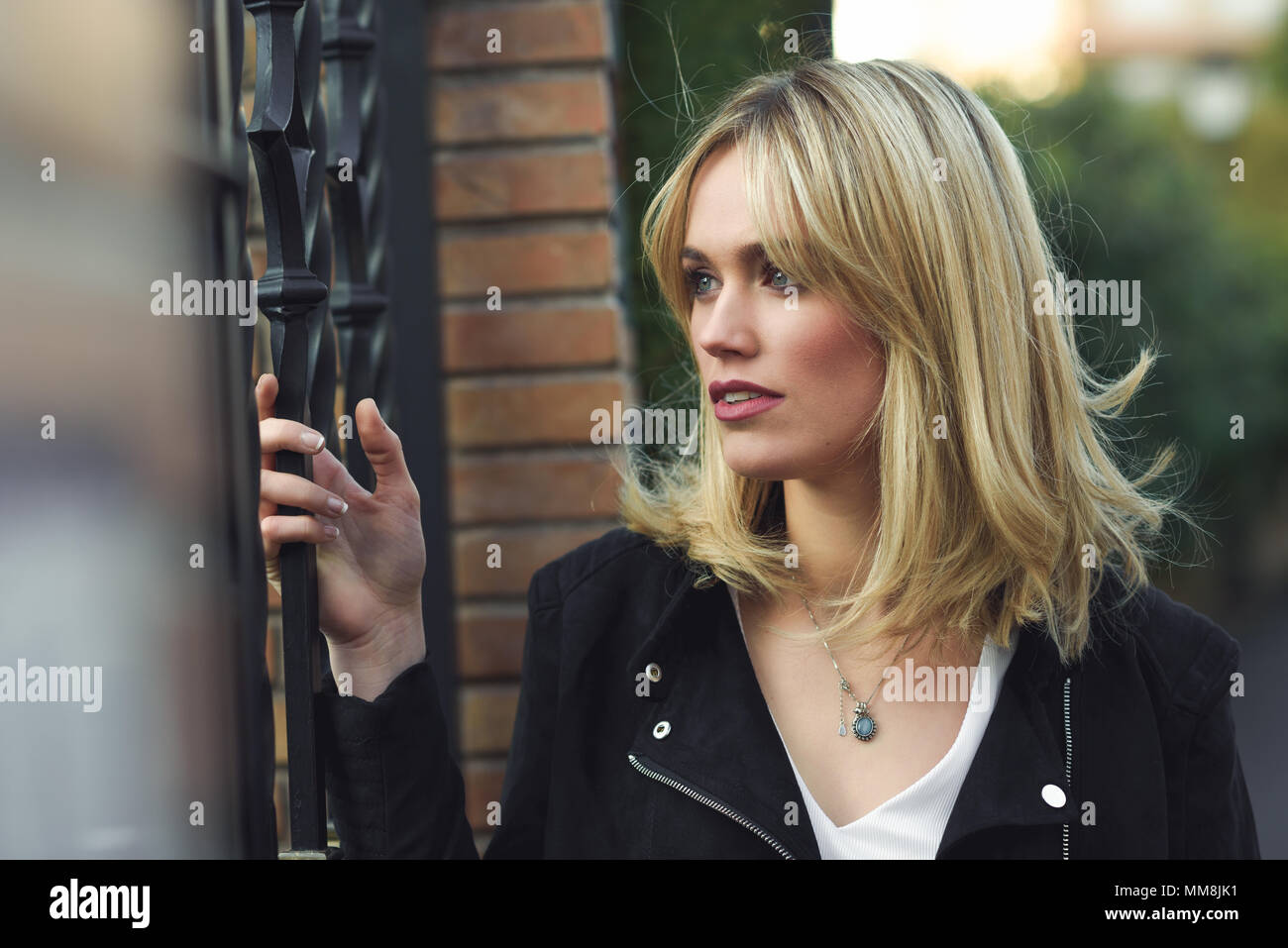 Attractive blonde woman standing in urban background with lost look. Young girl wearing black zipper jacket. Pretty female with straight hair hairstyl Stock Photo