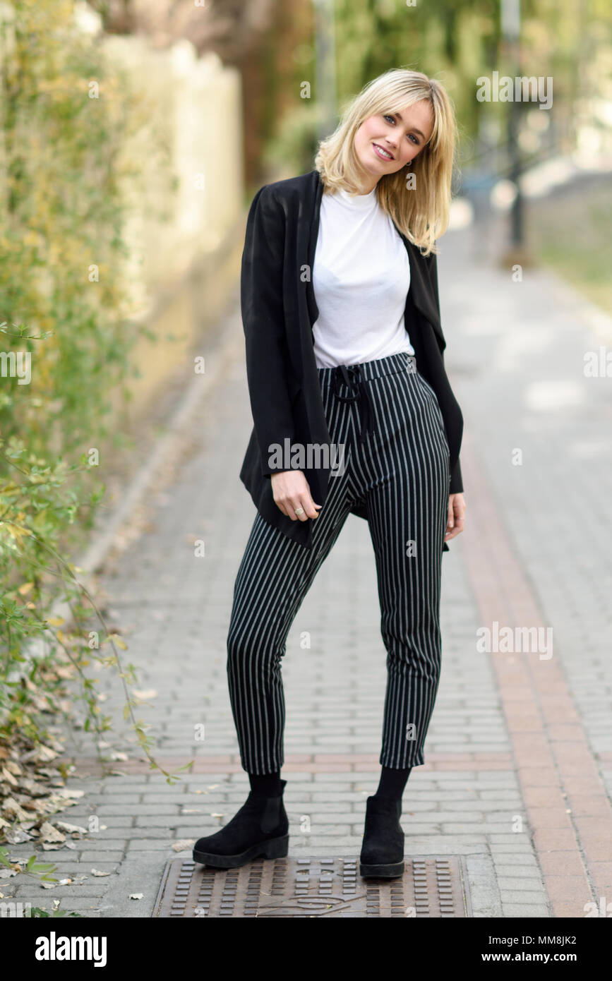 Pretty blonde woman smiling in urban background. Young girl wearing black blazer jacket and striped trousers standing in the street. Pretty female wit Stock Photo