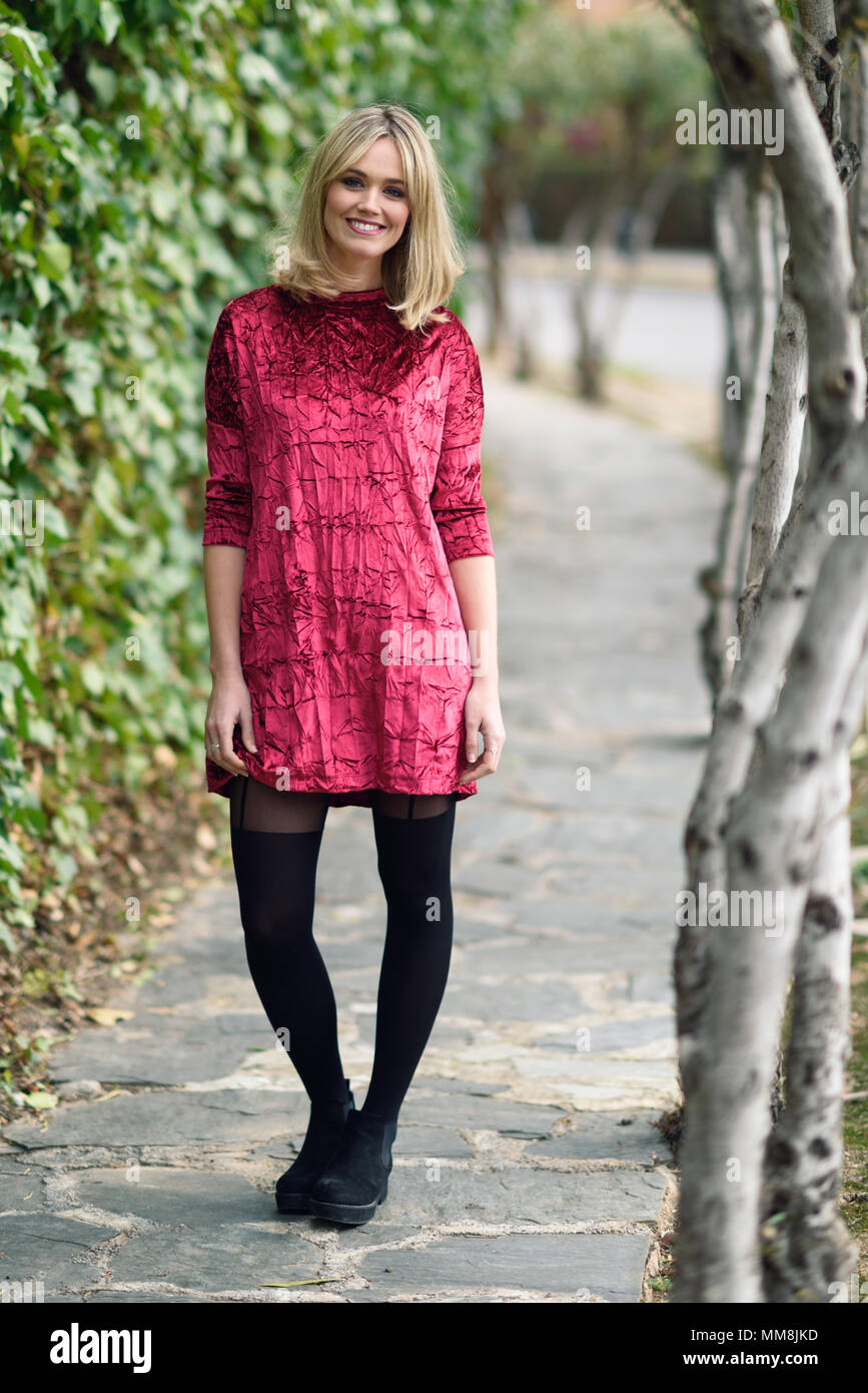 Beautiful blonde woman smiling in urban background. Young girl wearing red dress and tights standing in the street. Pretty female with straight hairst Stock Photo