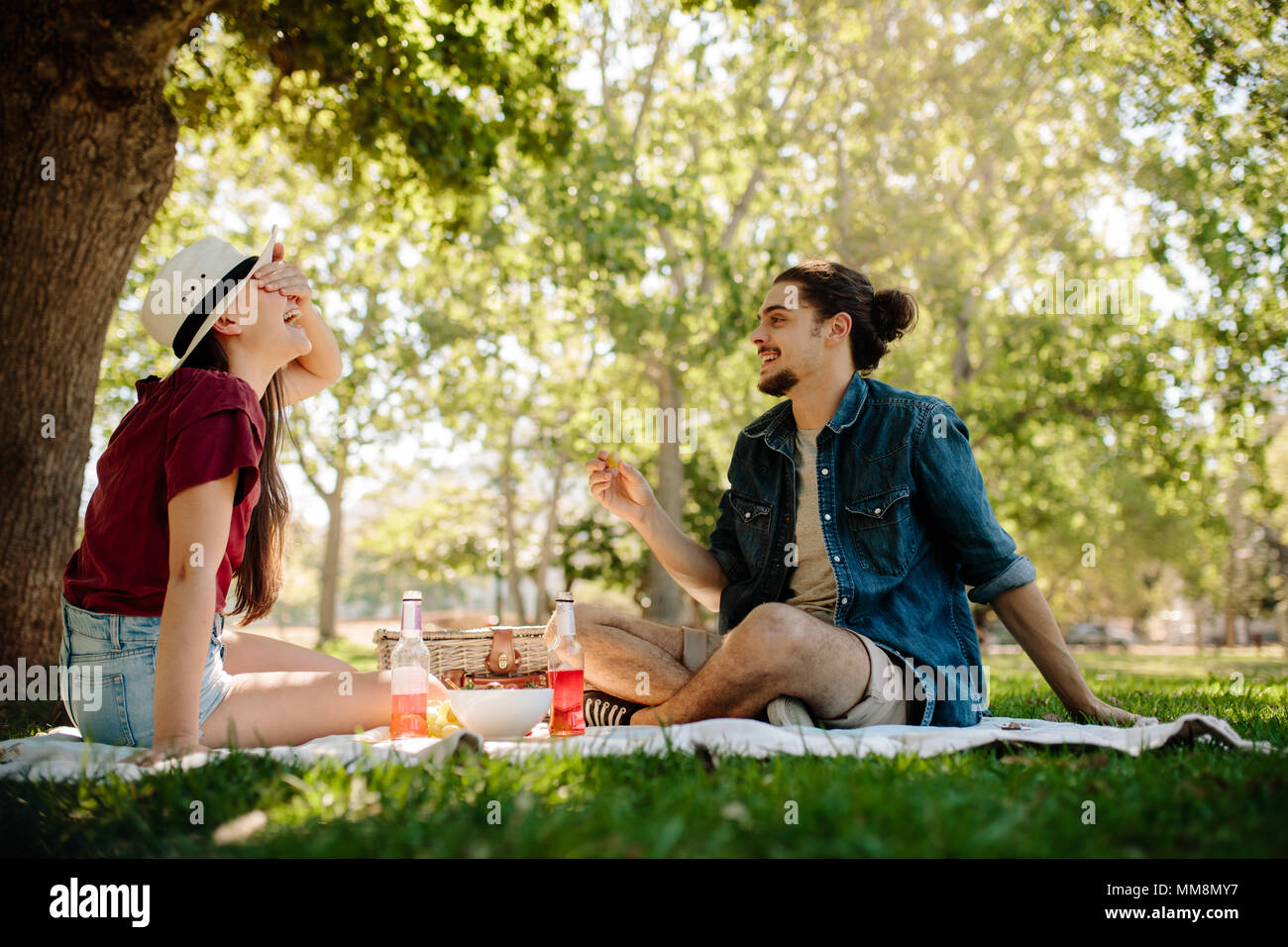 Couple playing a game on picnic at park. Woman covering her eyes with man holding a fruit in hand. Stock Photo