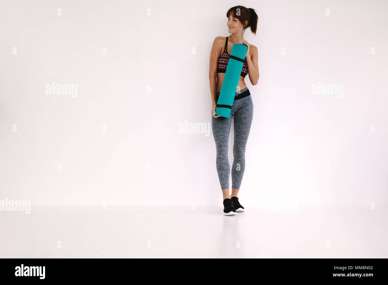 Full length of young sporty woman standing by a wall with yoga mat in studio. Caucasian female relaxing after yoga class. Stock Photo