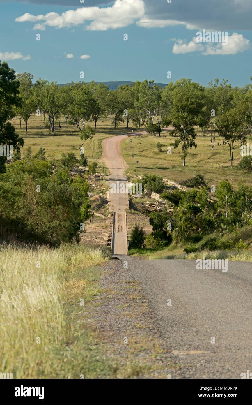 Steep road leading down to narrow low bridge across Bowen River & through landscape of trees & golden grasses under blue sky in Queensland Australia Stock Photo