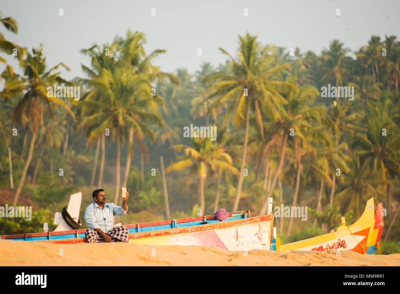 VARKALA - KERALA - INDIA - 31 JANUARY 2018. A man sitting on a wooden boat is taking a selfie on the beach of Varkala at Sunset. Kerala state, India. Stock Photo
