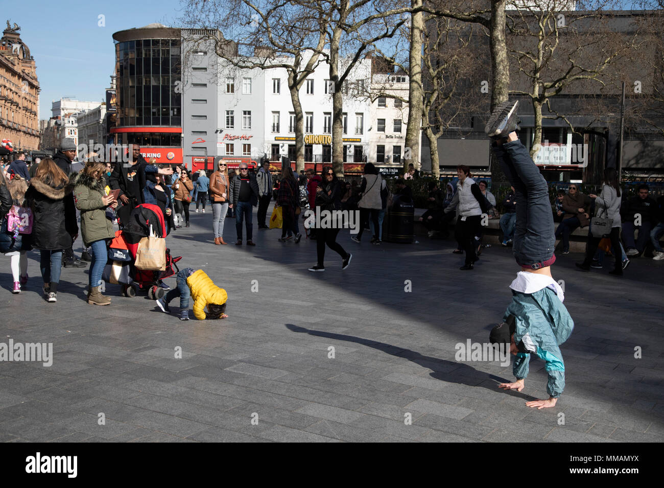 Young boy tries to mimick a street performer in Leicester Square who is performing a hand stand in London, England, United Kingdom. Stock Photo