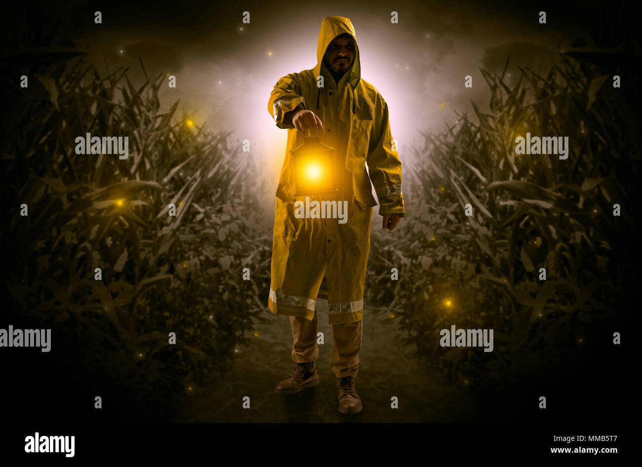 Man in raincoat at night coming from thicket and looking something with glowing lantern  Stock Photo