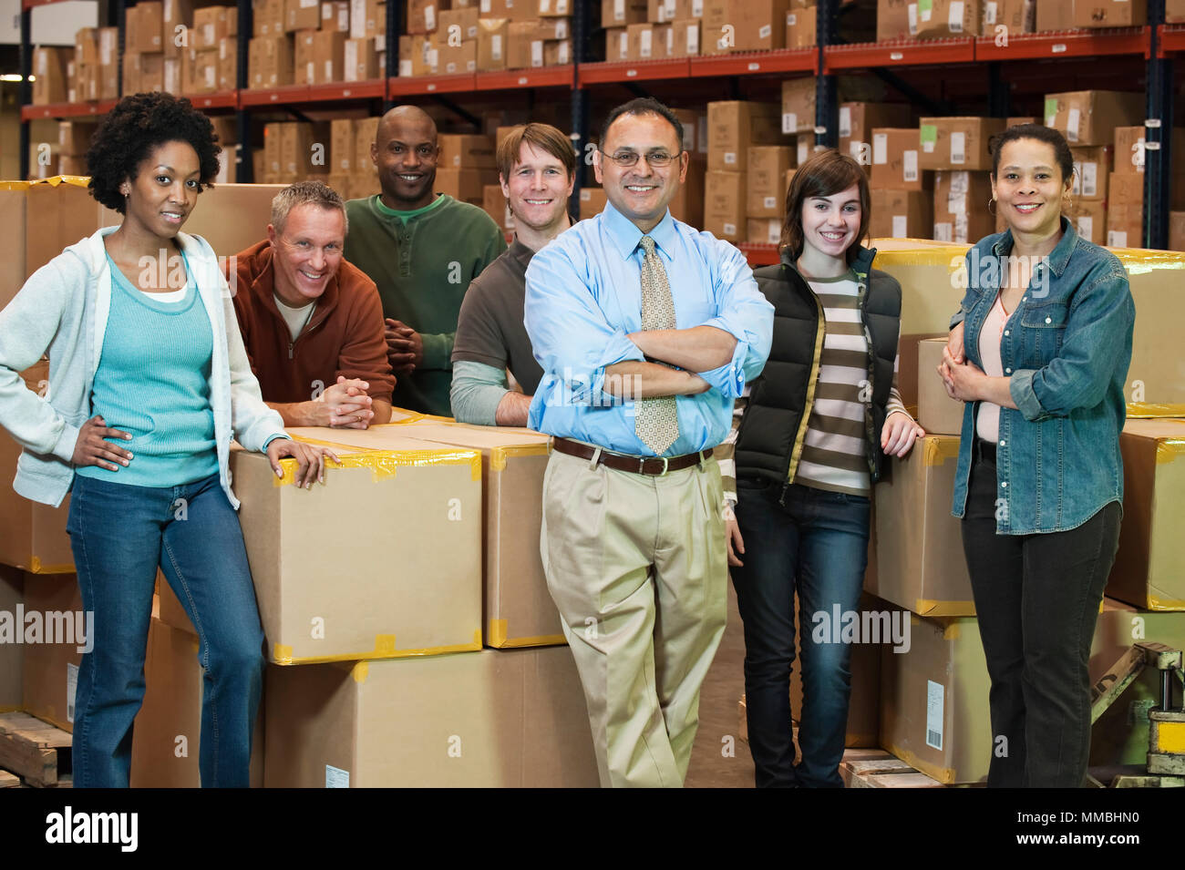 Team portrait of multi-ethnic  warehouse workers lead by a Hispanic American male executive and surrounded by large racks of products stored in cardbo Stock Photo