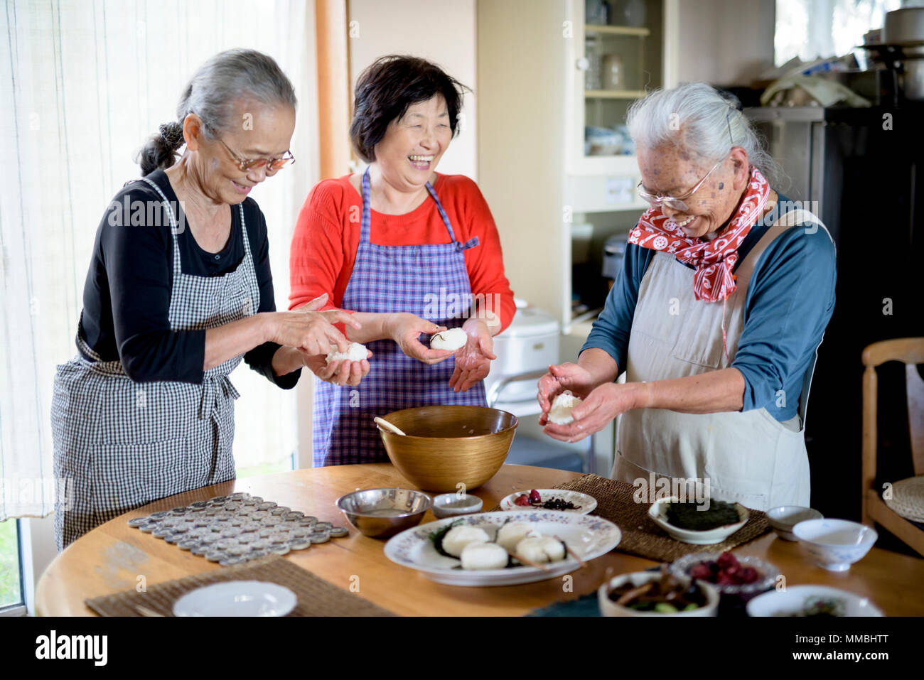 Three older women standing round a table in a kitchen, making sushi. Stock Photo
