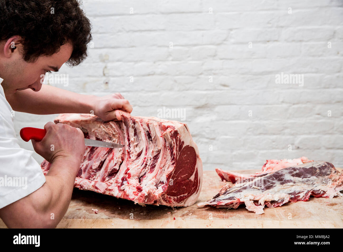 Close up of man wearing apron standing at a wooden butcher's block, butchering beef forerib. Stock Photo