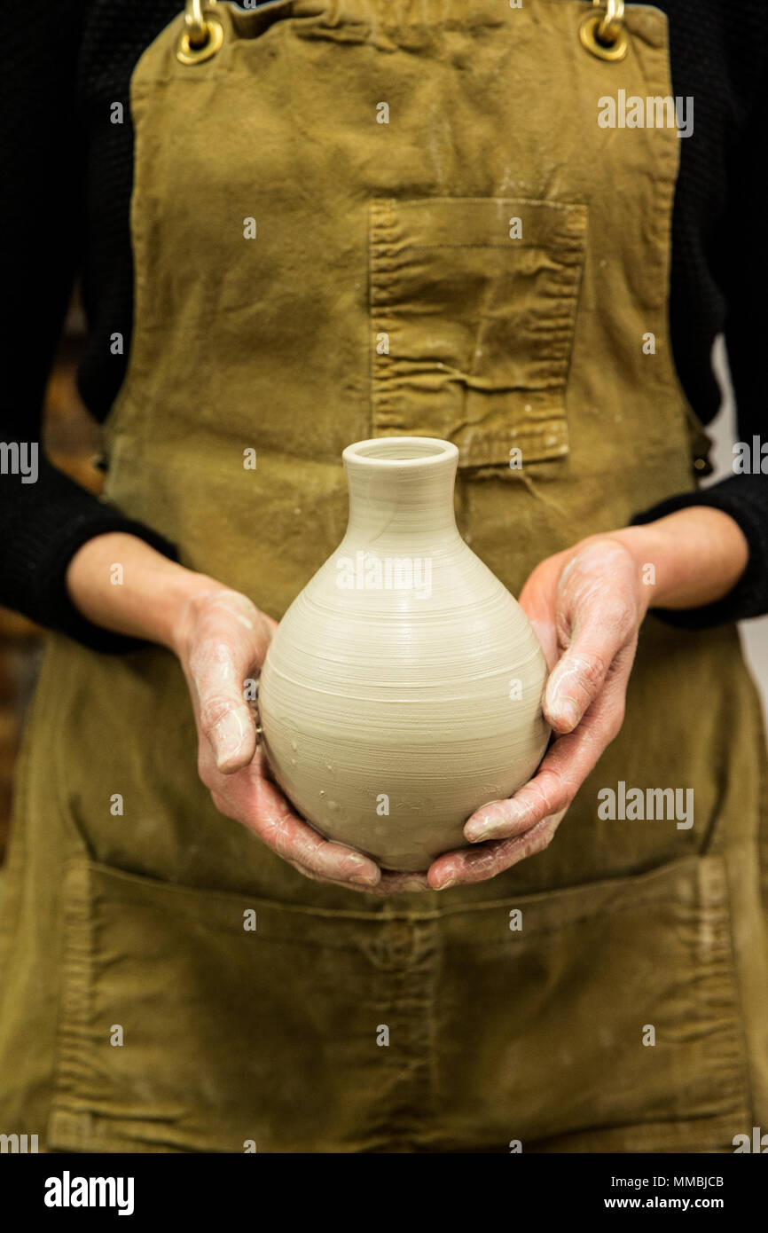 Close up of potter wearing apron holding unfired spherical clay vase ...