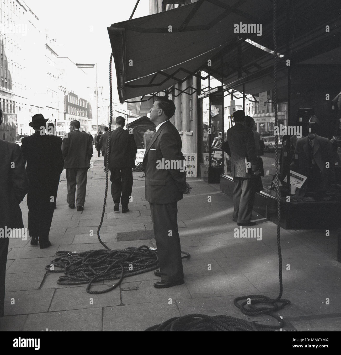 London, 1961, The Strand and a gentleman standing on the wide pavement in the shade underneath a shop's canopy or awning with rope lying on the ground indicating building work above. Stock Photo
