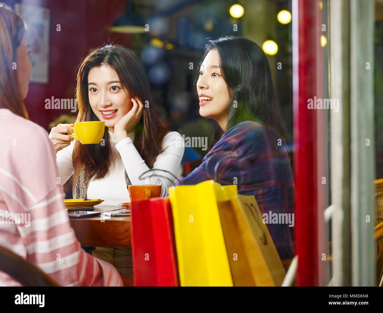 three happy beautiful young asian women sitting at table chatting talking in coffee shop or tea house after shopping, shot through window glass. Stock Photo