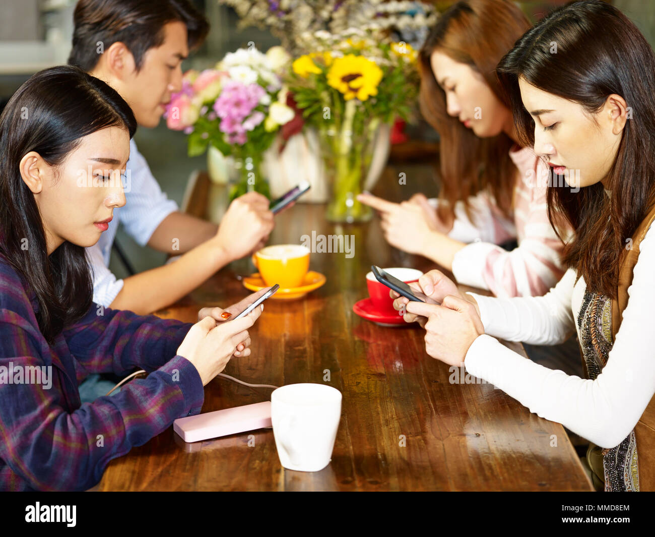 four young asian adults man and women sitting in coffee shop playing with mobile phone with very serious facial expression. Stock Photo