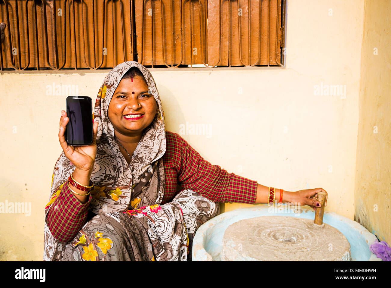 Rural Female Using Grind Stone Mill .And Showing Cell-Phone At-Home Stock Photo