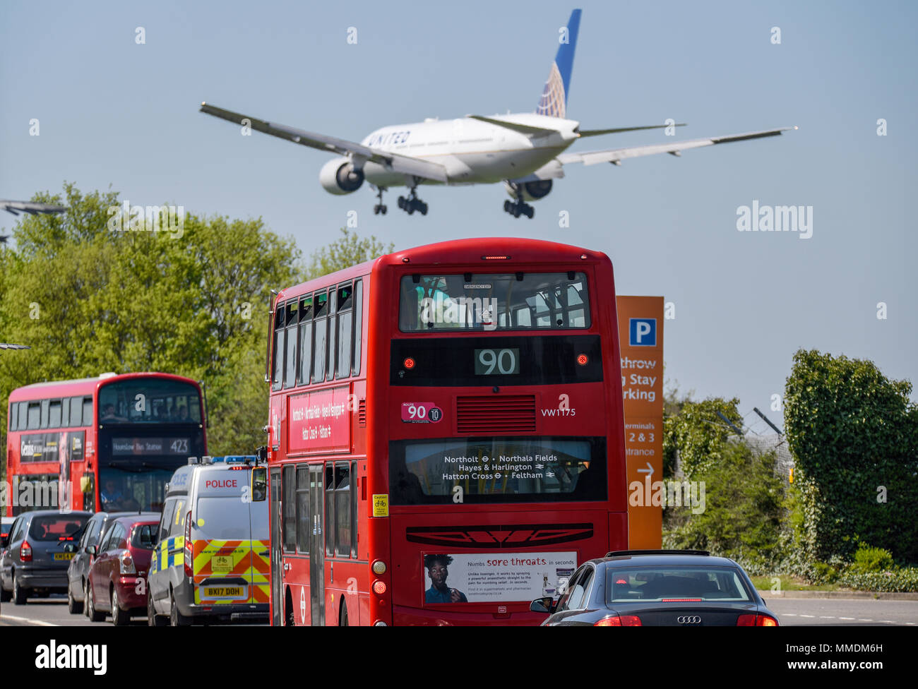 United Airlines Boeing 777 jet plane coming in to land at London Heathrow Airport, UK, in blue sky, over a red London bus, police vehicle Stock Photo