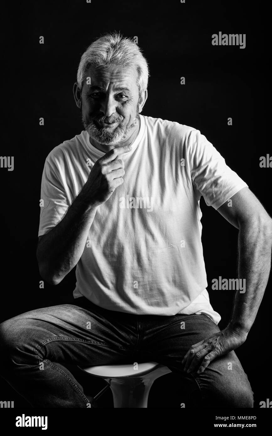 Mature man smiling looking at camera. Senior male with white hair and beard laughing wearing casual clothes isolated on black background. Studio shot  Stock Photo