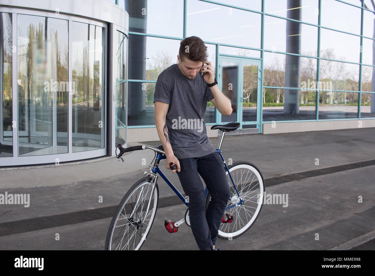 young hipster use cellphone, fixed gear bike on city street. big mirror windows background, urban bicycle rider Stock Photo