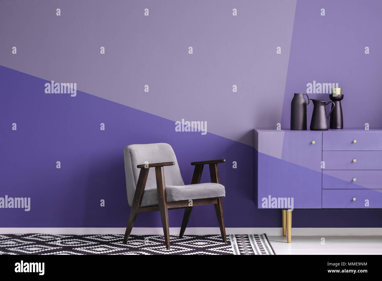 Real photo of a gray, wooden armchair on patterned, black and white rug in creative living room interior with geometric, violet wall and cupboard Stock Photo