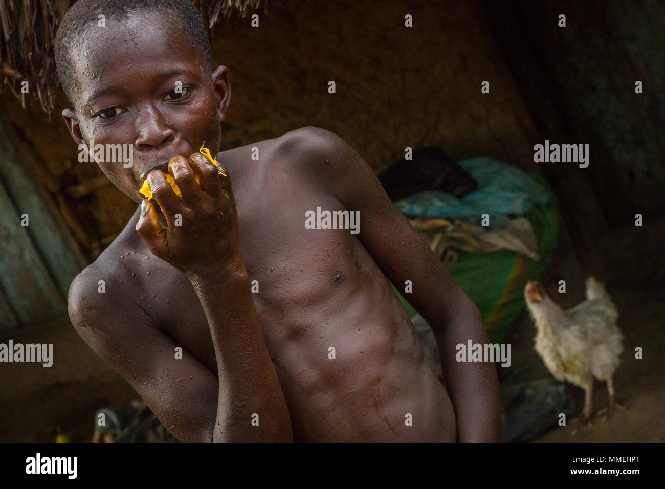 YONGORO, SIERRA LEONE - June 06, 2013: West Africa, portrait of an unknown boy while eating a yellow fruit in the background a chicken, near the capit Stock Photo