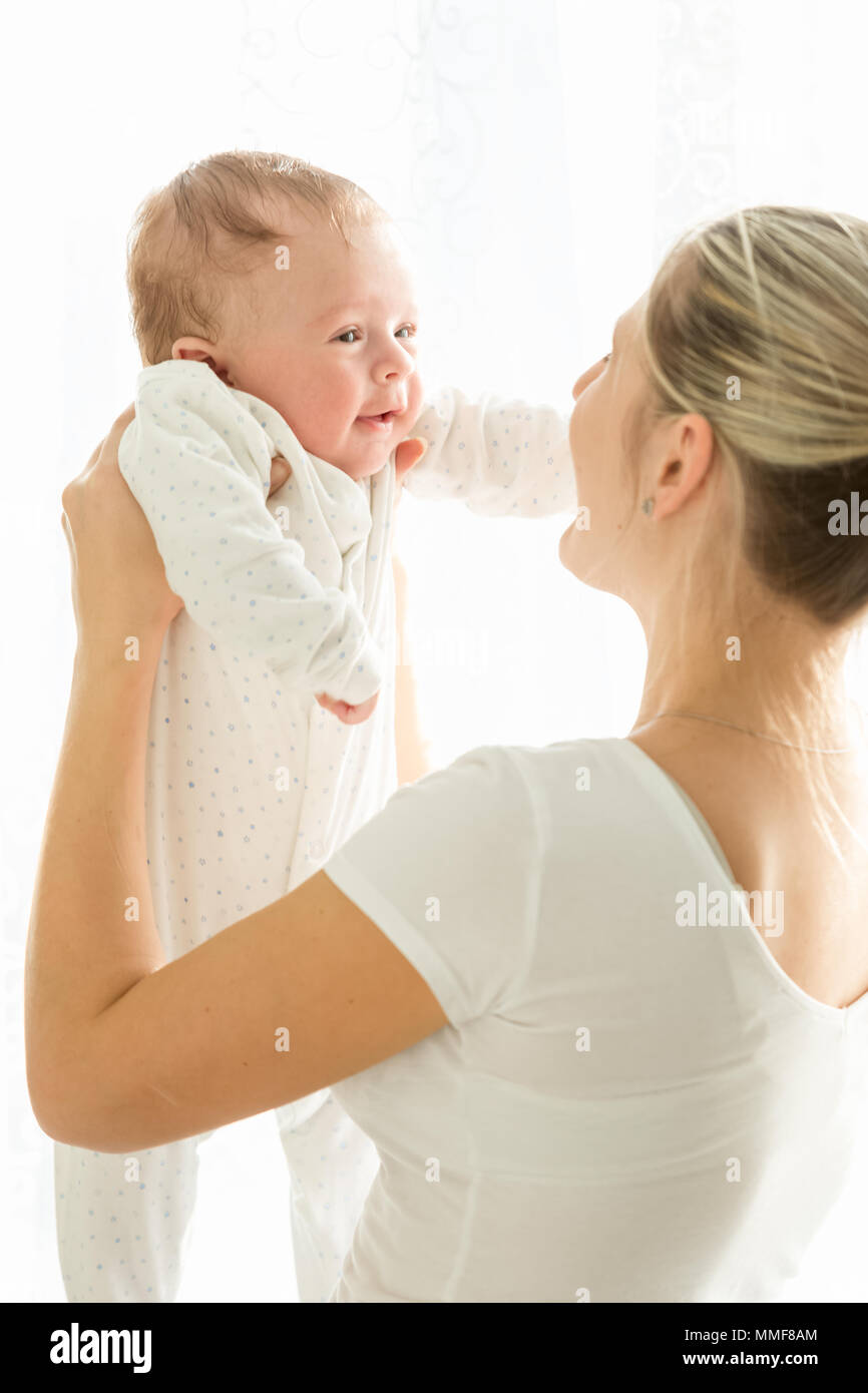 Happy young woman holding her smiling 3months old baby on stretched hands at window Stock Photo