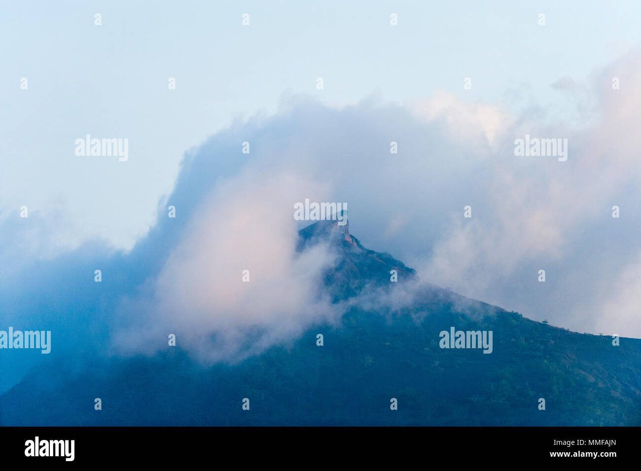 Mist shrouds the jagged summit of a tropical island covered in rainforest. Stock Photo