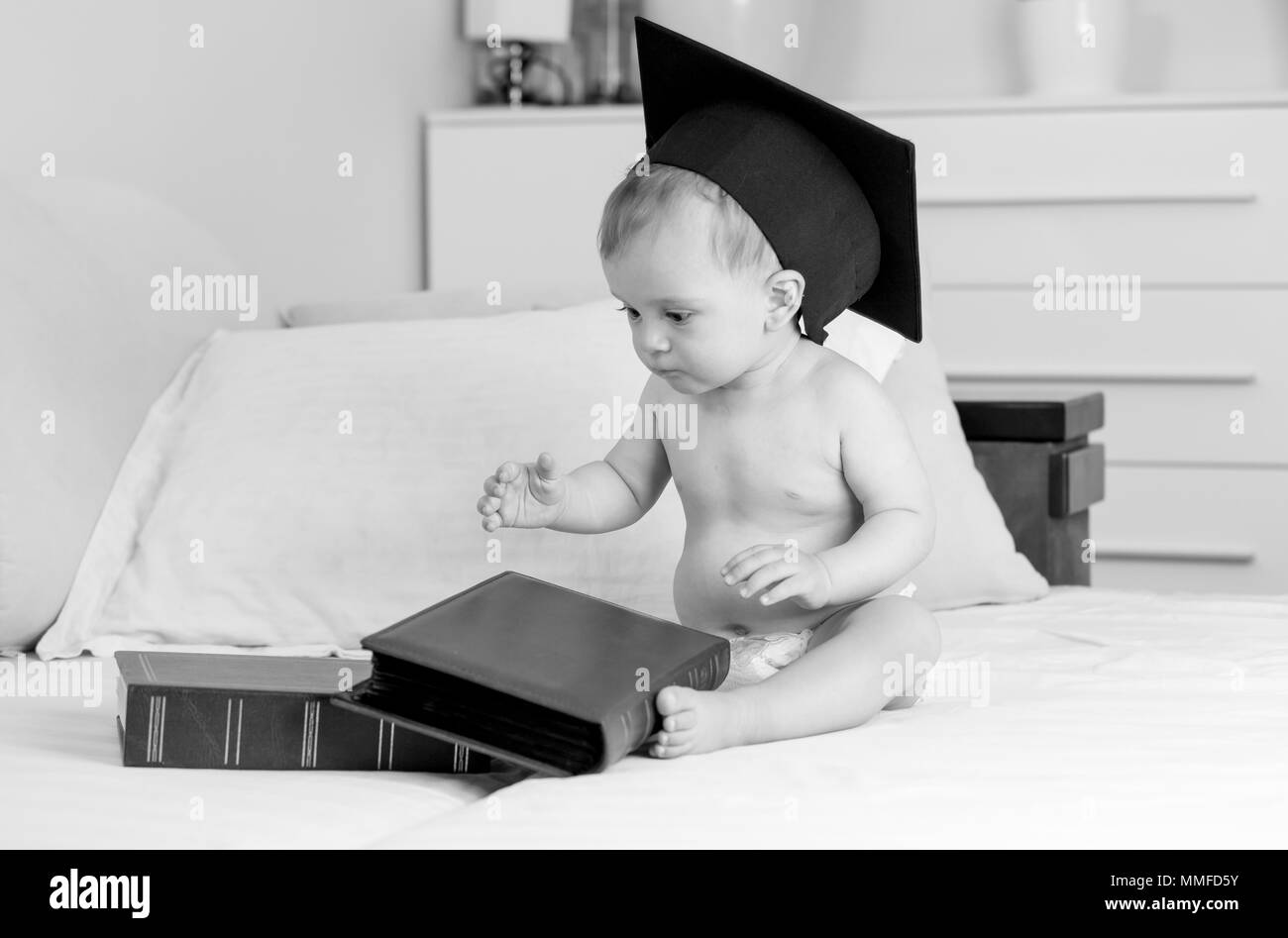 Black and white photo of baby boy in graduation cap sitting on bed and looking on big pile of books Stock Photo