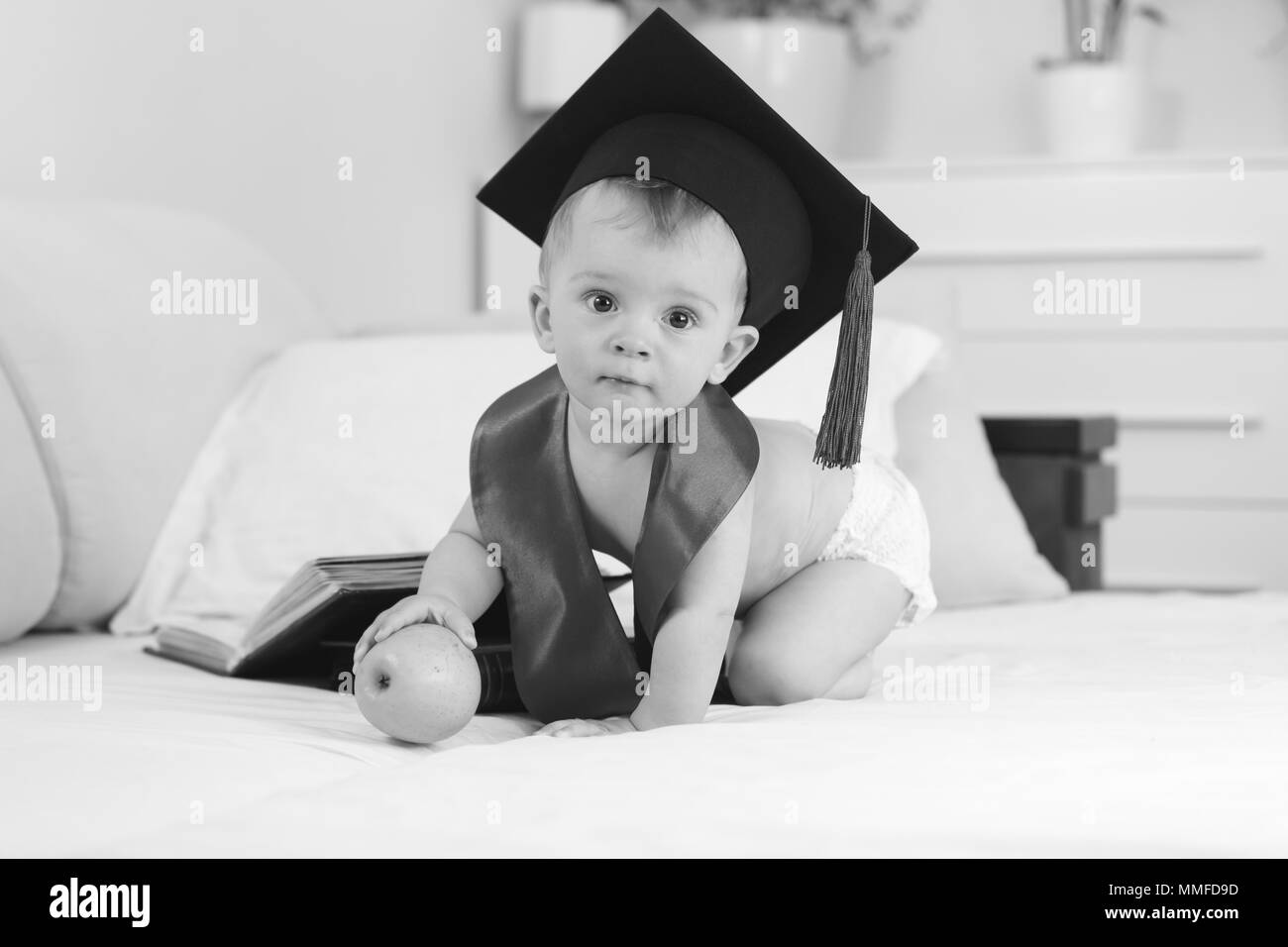 Black and white image of genius boy in graduation hat sitting on bed with apple and looking in camera Stock Photo