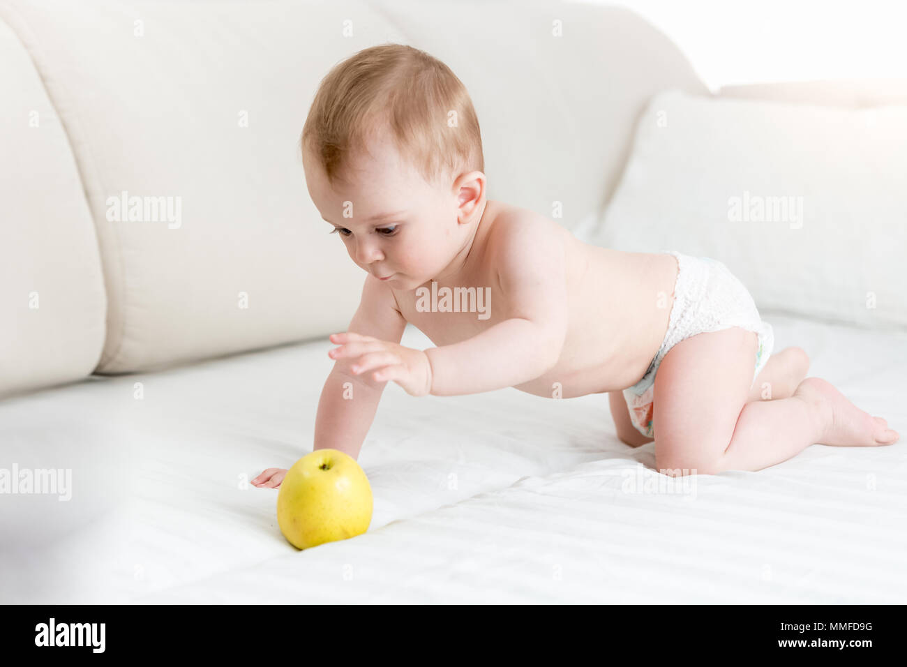 Cute baby boy in diapers crawling on bed and reaching for big yellow