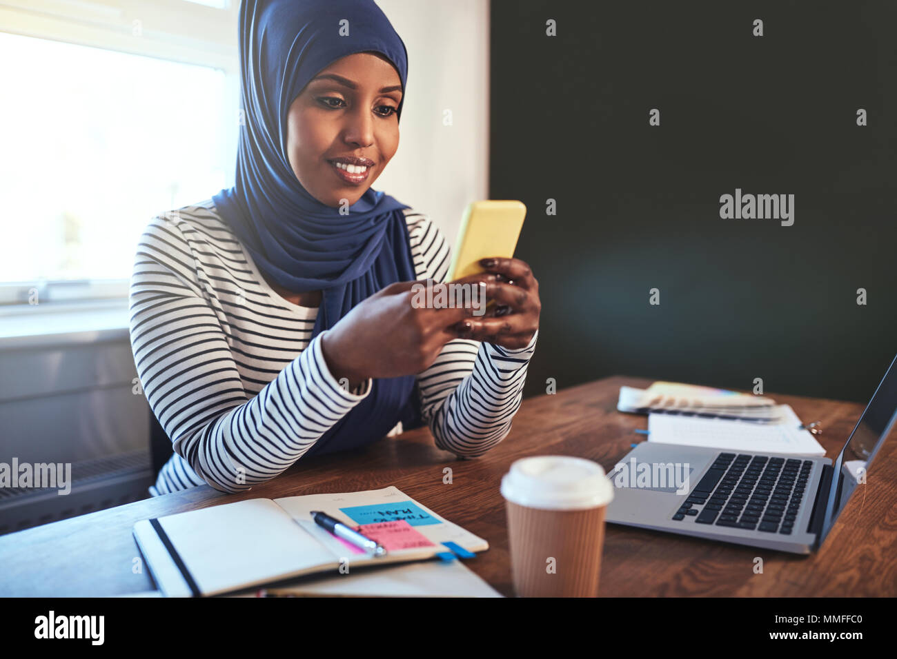 Smiling young Arabic female entrepreneur wearing a hijab sending a text ...