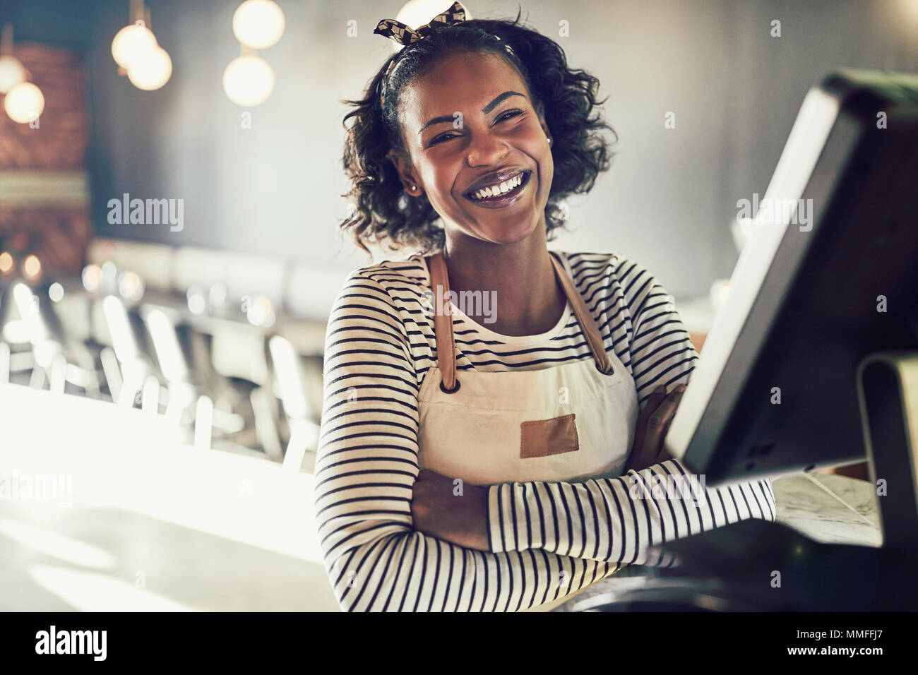 Young African waitress wearing an apron and laughing while standing by a point of sale terminal in a trendy restaurant Stock Photo
