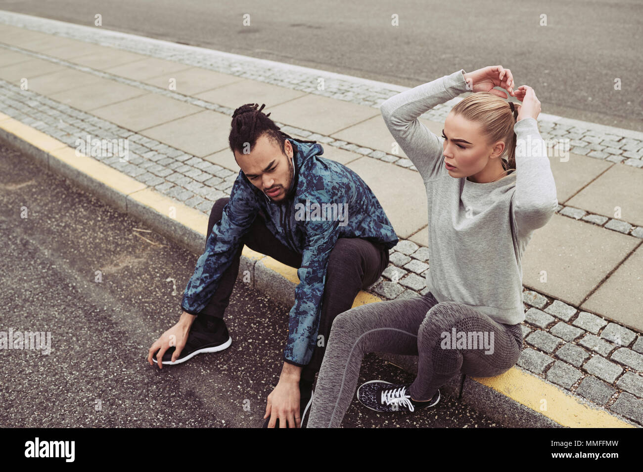 Diverse young couple in sportswear sitting by a road and catching their breath while jogging together outdoors Stock Photo