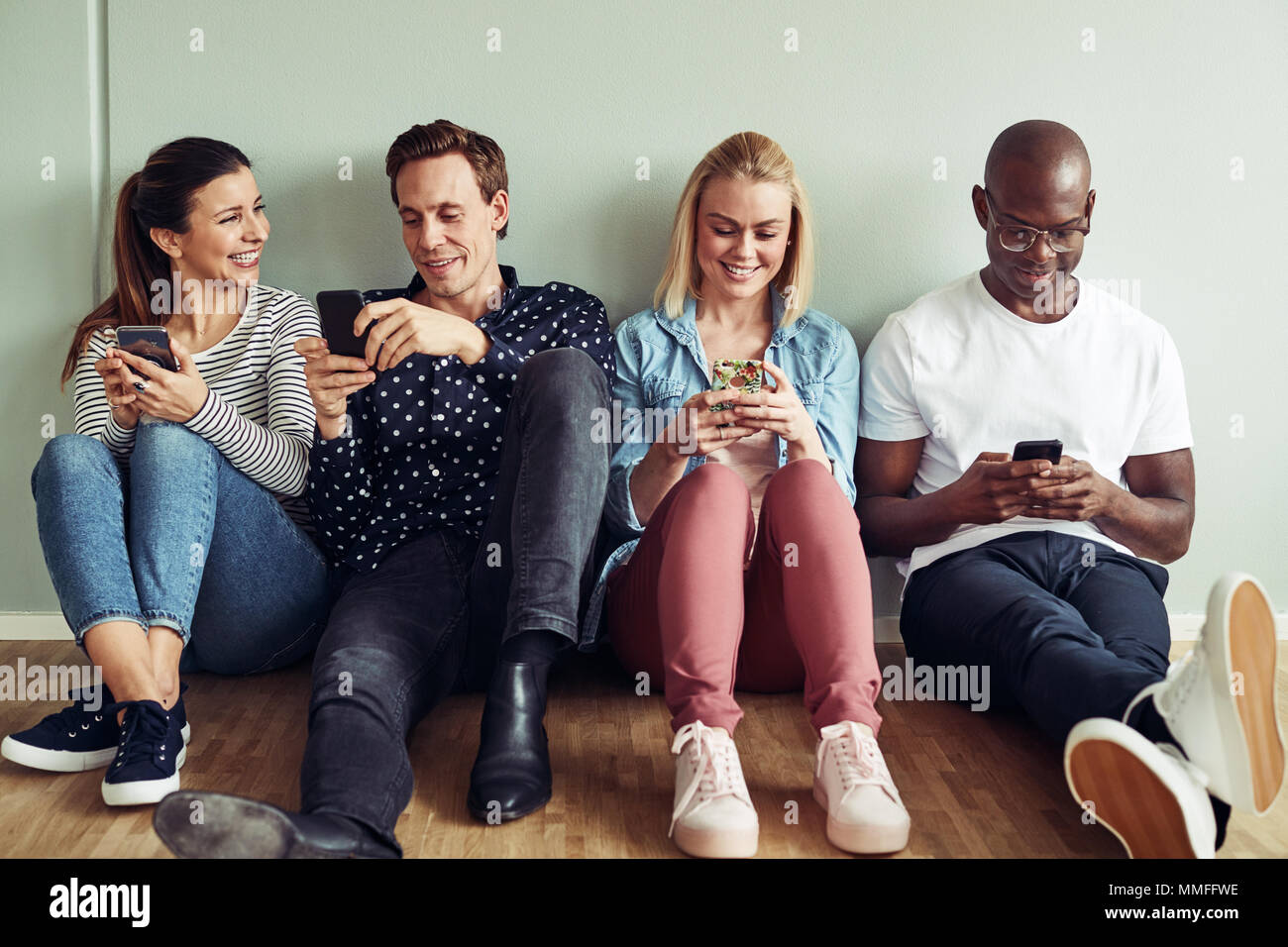 Smiling group of diverse young coworkers sitting together on the floor of an office using their cellphones during a break Stock Photo