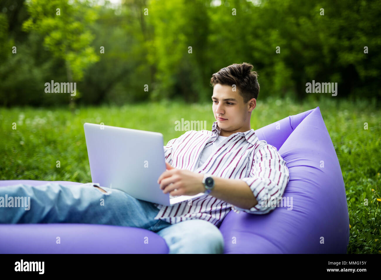 Handsome man lying on inflatable sofa working on laptop while resting on grass in park Stock Photo