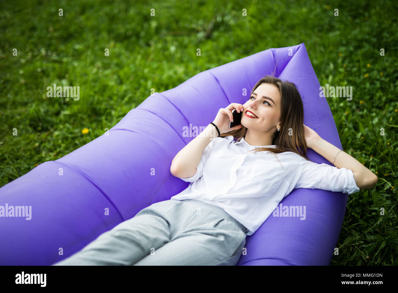 Pretty young woman lying on inflatable sofa lamzak talk on phone while resting on grass in park on the sun Stock Photo