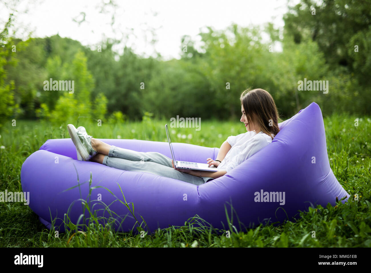 Pretty young woman lying on inflatable sofa lamzak working on laptop while resting on grass in park on the sun Stock Photo