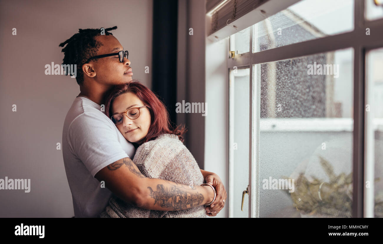 Interracial couple hugging each other while standing near window. African man embracing his caucasian girlfriend at home. Stock Photo