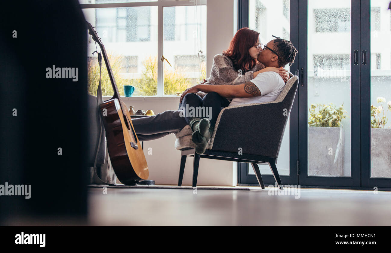Interracial couple sitting on a armchair and kiss one another. Woman sitting on man's lap and kissing. Stock Photo