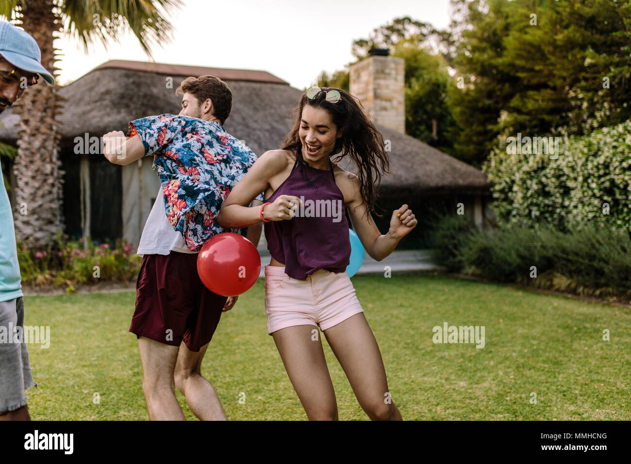Woman trying to pop a balloon with her hips tied on man's back. Friends playing balloon popping games at party outdoors. Stock Photo