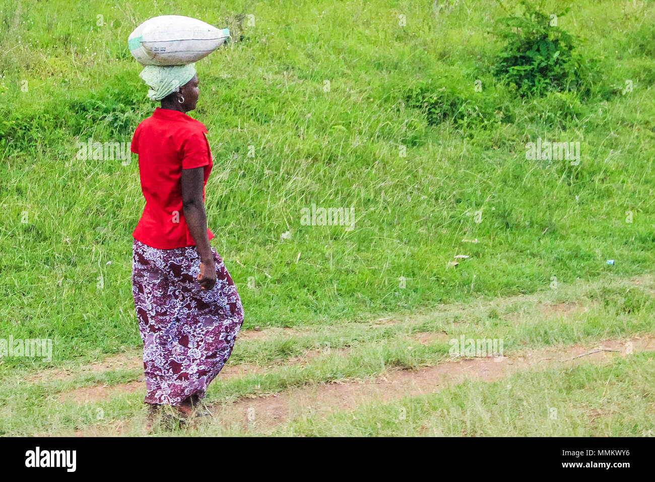 Arusha, Tanzania, Africa - January 12, 2013:  A young woman walks carrying an heavy bag on her head for a long distance to home. Lack of transports in the area, force the people to walk a lot on the road to move from town to home Stock Photo