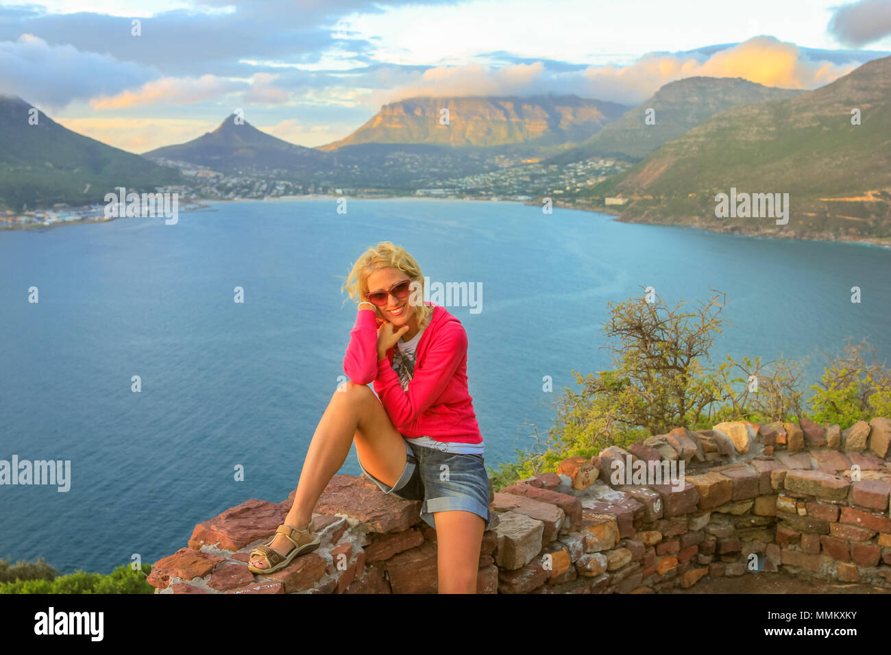 Happy woman enjoying aerial view of Sentinel peak in Hout Bay from Lookout Point in Hout Bay on Chapman's Peak Drive, Cape Town, South Africa. Lifestyle woman traveler in day trip road. Twilight shot. Stock Photo