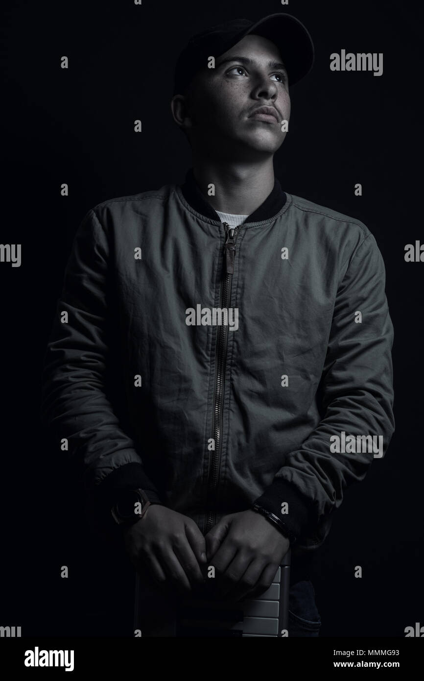teen boy posing and looking up with black cap and bomber jacket in front of black background Stock Photo