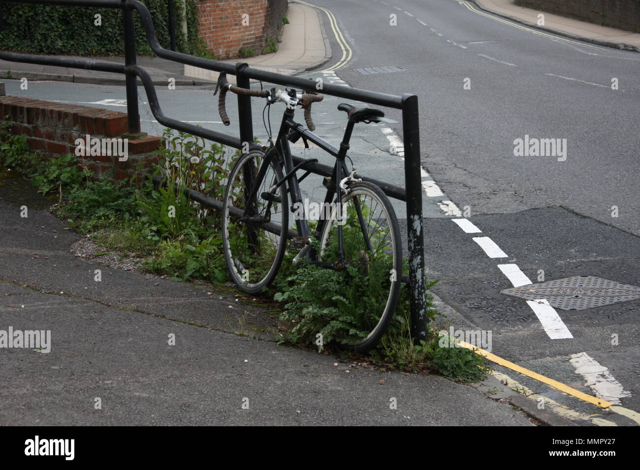 A bike leans, lonely against the side of the road, weeds poking up through its spokes in a snapshot of urban life. Stock Photo