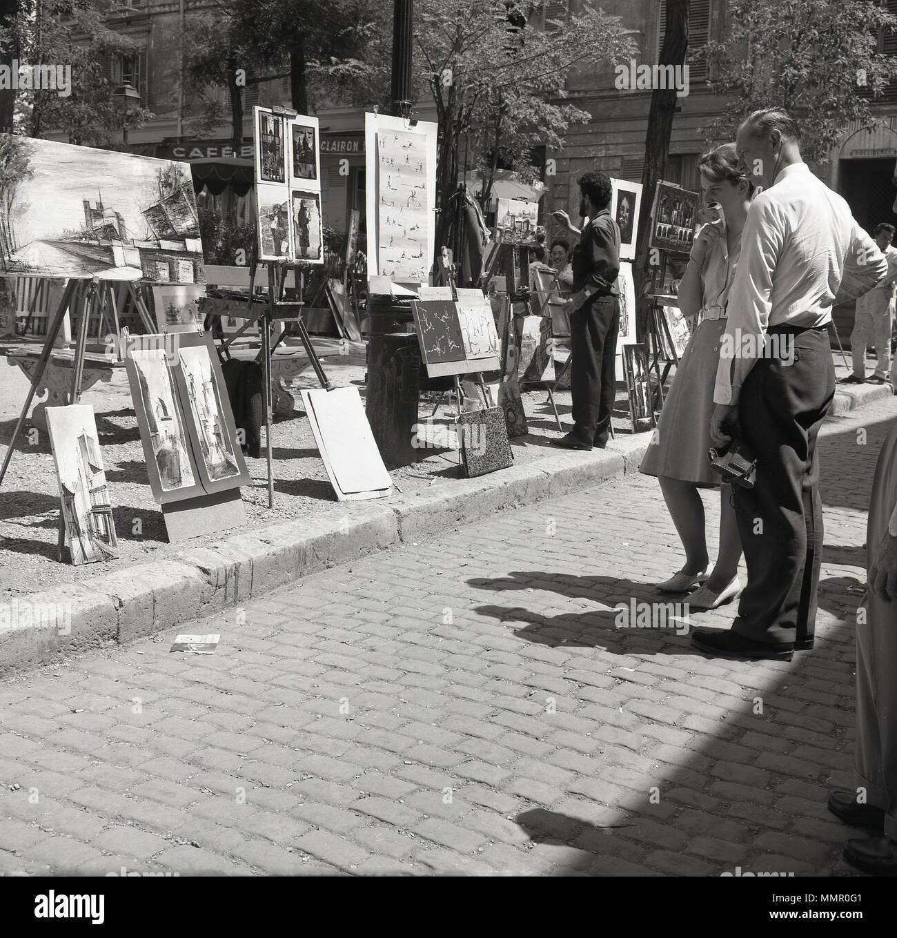 1950s, couple standing looking at the artwork displayed on the square of Place du Tertre at Montmartre, Paris, France. The area, in the northern part of the city, is famous for its artistic history and street artists use the main square to create and display their work. Stock Photo